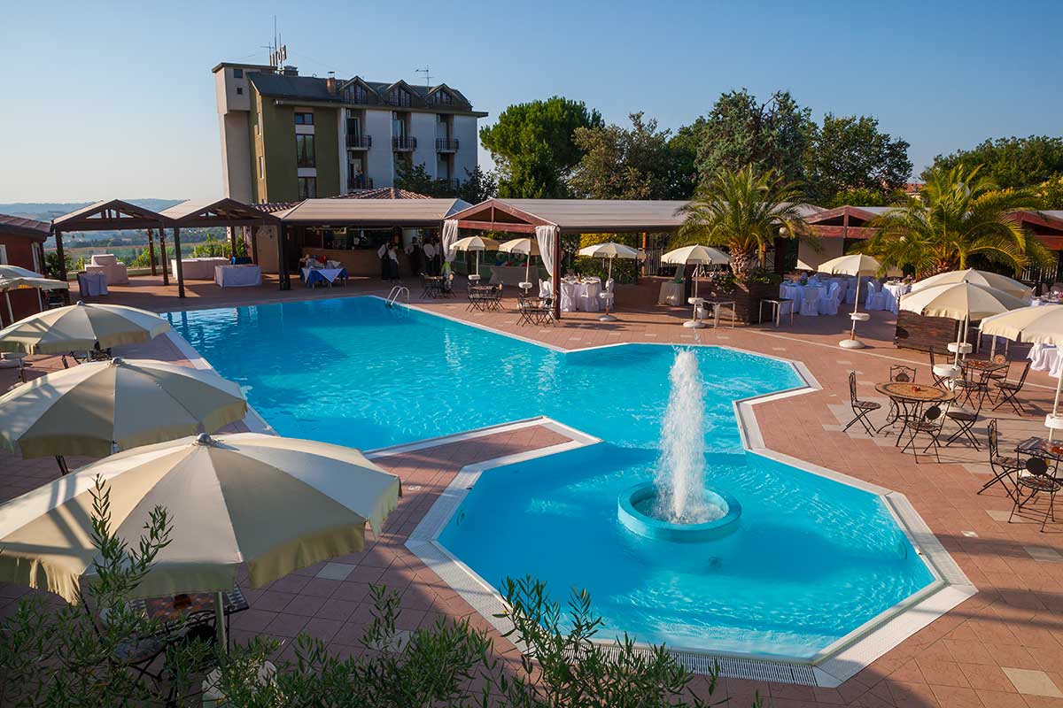 Piscina all'aperto con area eventi attrezzata con tavoli rotondi e sedie coperte da tovaglie bianche, ombrelloni e palme